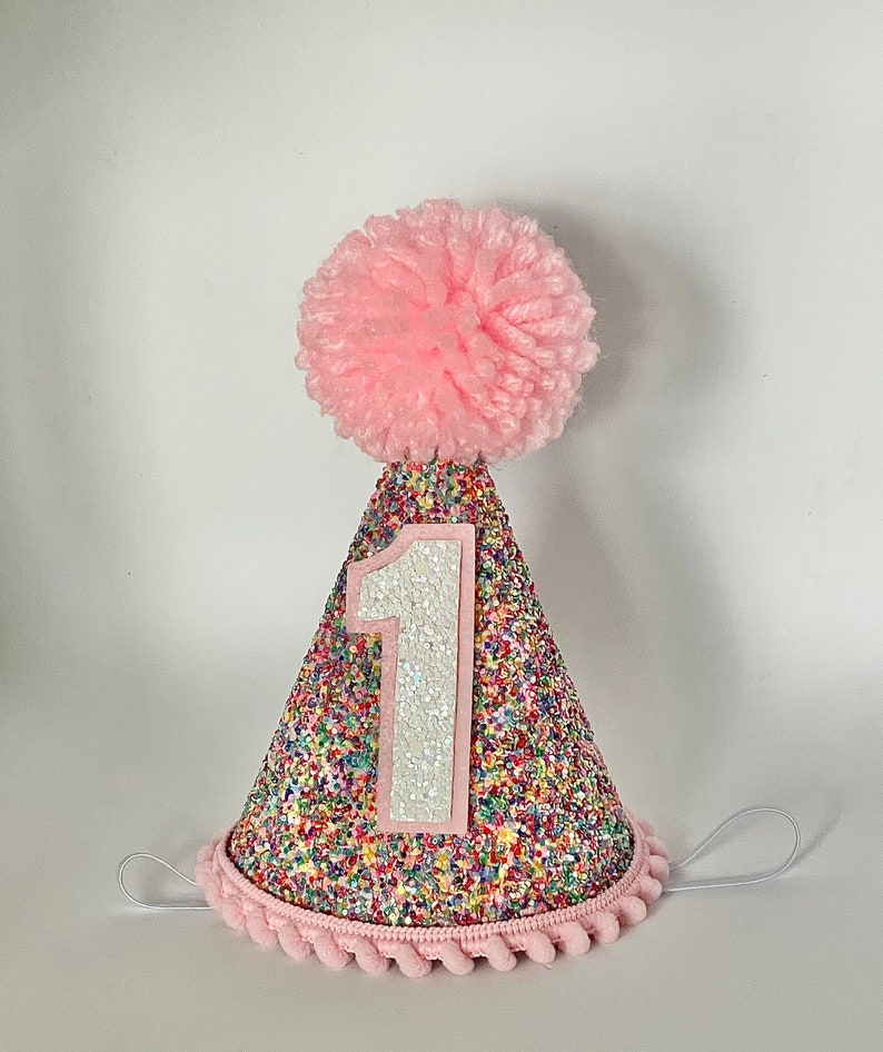 May include: Pink and rainbow glitter party hat with a white number one and a pink pom pom on top.