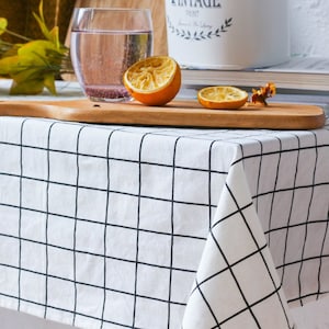 Puede incluir: Un mantel blanco con un patrón de cuadrícula negra cubre una mesa. Una tabla de cortar de madera sostiene una naranja cortada por la mitad y una rodaja. Un vaso de agua y una lata de pintura vintage están en el fondo.