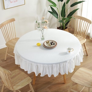 May include: White round tablecloth with a ruffled edge, set on a wooden table with four chairs. The tablecloth has a delicate pattern and is decorated with a small plate of fruit, a cup, and a vase of white flowers.