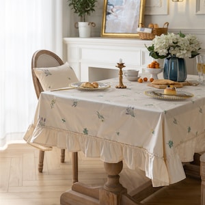 May include: A white tablecloth with a floral pattern and a ruffled edge. The tablecloth is laid on a wooden table with a wooden base. There are two plates with pastries on the table, a candle holder, and a vase with flowers.