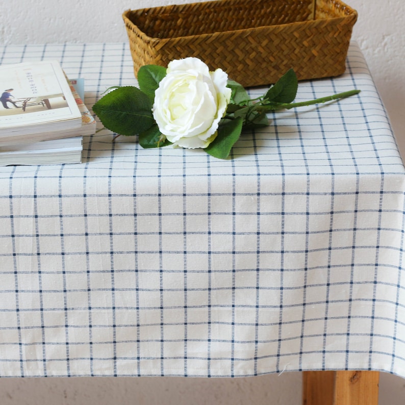 May include: A white tablecloth with a blue checkered pattern. A white rose with green leaves is on the tablecloth. A woven basket is in the background.