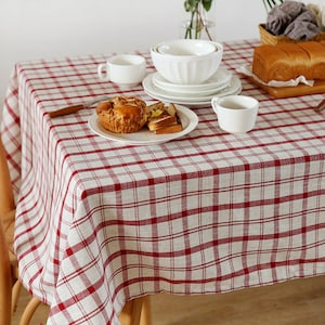 Può includere: Una tovaglia a quadri rossi e bianchi con una ciotola di ceramica bianca, due tazze di ceramica bianche, un piatto di pasticcini e un tagliere di legno con del pane sopra.