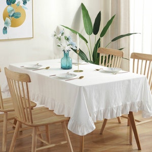 Puede incluir: Mantel blanco con borde de volantes, colocado sobre una mesa de madera con cuatro sillas. Un jarrón de vidrio azul con flores blancas y un candelero dorado están sobre la mesa.