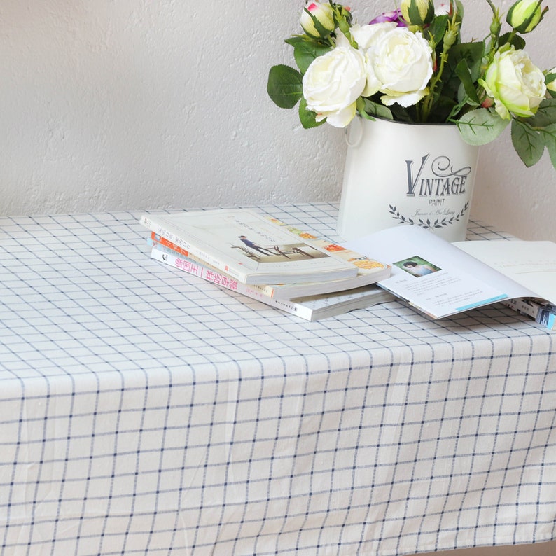 May include: A white tablecloth with a blue and white checkered pattern. The tablecloth is on a table with a stack of books and a vase of white flowers. The vase is white with the text "Vintage Paint" on it.