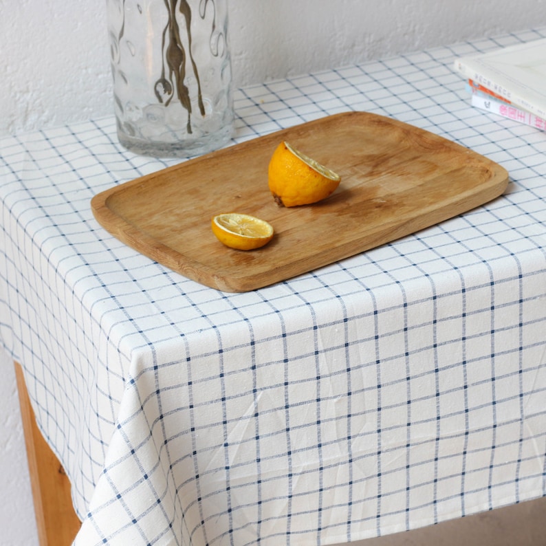 May include: A white tablecloth with a blue checkered pattern, a wooden serving tray with two lemon halves on top, and a clear glass vase with dried flowers.