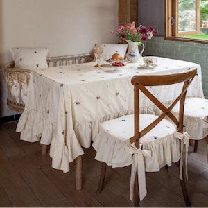 May include: A white tablecloth with a floral pattern and ruffled edges covers a wooden table with two chairs. The chairs have white cushions with ruffled edges and bows. The table is set with a vase of flowers, a plate of fruit, and a cup and saucer.