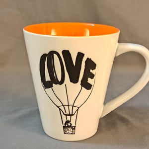 May include: White ceramic mug with an orange interior. The mug features a black and white illustration of a hot air balloon with the word "LOVE" written in the balloon.