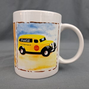 May include: White ceramic mug featuring a yellow Coca-Cola delivery truck with black wheels and the Coca-Cola logo. The mug has a handle and a decorative border with a blue sky and sandy ground background.