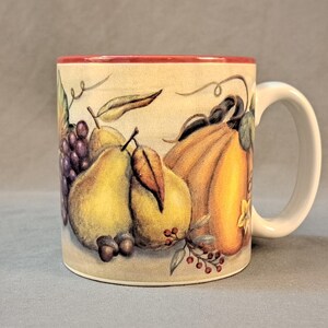 May include: A white ceramic mug with a red rim and a colorful autumn harvest design featuring pears, grapes, and a pumpkin. The design is painted on the mug and includes a brown vine and red berries.