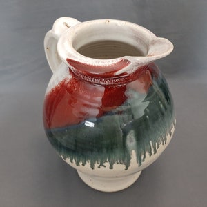 May include: A white ceramic pitcher with a red and green glaze. The pitcher has a round body and a wide mouth.