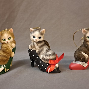 May include: Three decorative cat figurines perched in high-heeled shoes. The cats are in various colors, including orange, gray, and brown. The shoes are green, black with white polka dots, and red. Each cat has a gold string for hanging.