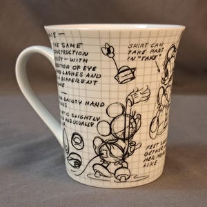 Op de afbeelding: Witte keramische mok met een zwart-witte illustratie van Mickey Mouse en Minnie Mouse. De mok heeft een handvat en bevat tekst over het ontwerp van de personages.