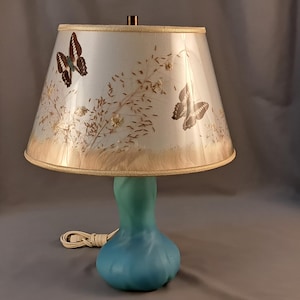 May include: A teal blue ceramic table lamp with a white lampshade featuring a butterfly and floral design.