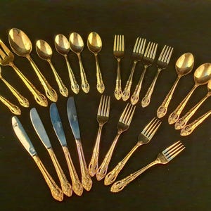 May include: A collection of gold-colored silverware, including forks, spoons, and knives, arranged on a dark surface. The silverware has ornate detailing on the handles, suggesting a vintage or antique style. The arrangement showcases a complete set for dining.
