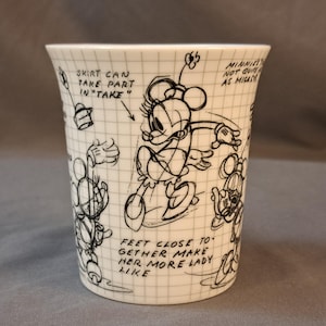 Op de afbeelding: Een witte keramische mok met een zwart-witte illustratie van een cartoonfiguur. De illustratie toont een vrouwelijk figuur met een rok, en de tekst "SKIRT CAN TAKE PART IN "TAKE"" staat op de mok. De tekst "MINNIE'S NOT QUITE AS MIGACH" staat ook op de mok. De tekst "FEET CLOSE TO GETHER MAKE HER MORE LADY LIKE" staat op de mok.