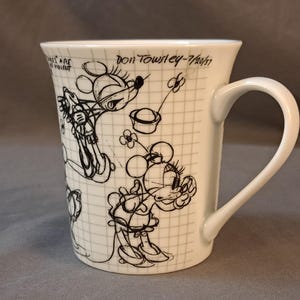 Op de afbeelding: Witte keramische mok met een zwart-witte schets van Mickey Mouse op een geruit achtergrond. De mok heeft een handvat en de tekst "Don Towley - 7/20/17" staat op de zijkant gedrukt.