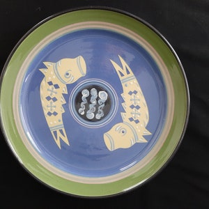 May include: A blue and green ceramic plate with a stylized design of two fish. The fish are yellow with blue and white details. The plate has a green rim and a white center.