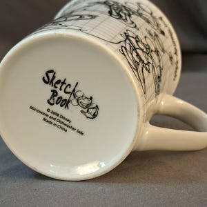 Op de afbeelding: Een witte keramische mok met een zwart-witte schets van Mickey Mouse en andere Disney-figuren. De onderkant van de mok heeft de tekst "Sketch Book" met het copyright "&copy; 2008 Disney" en "Microwave and Dishwasher Safe. Made in China."