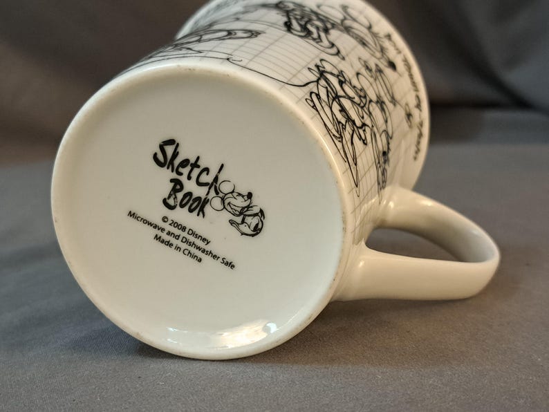 Op de afbeelding: Een witte keramische mok met een zwart-witte schets van Mickey Mouse en andere Disney-figuren. De onderkant van de mok heeft de tekst "Sketch Book" met het copyright "&copy; 2008 Disney" en "Microwave and Dishwasher Safe. Made in China."