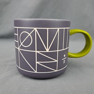 May include: A gray ceramic mug with a white geometric design and a green handle. The mug has a white interior and a small logo with the letters "R" on the side. The mug is approximately 4 inches tall.