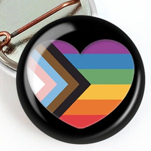 May include: A round, black button with a heart-shaped design featuring the colors of the Pride flag, including rainbow stripes and the Progress Pride flag. The button is next to a silver-colored pin back.