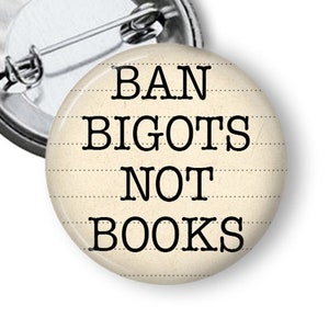 May include: A white button with black text that reads "BAN BIGOTS NOT BOOKS" on a lined background.