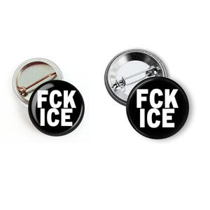 Puede incluir: Dos insignias redondas negras con el texto blanco "FCK ICE". Una insignia muestra la parte trasera del alfiler, la otra la parte delantera. Las insignias est&aacute;n sobre un fondo blanco.