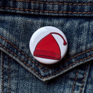 May include: A round, white button pin featuring a cartoon illustration of a red beanie hat with a tassel. The pin is set against a blue denim jacket.