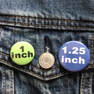 Peut inclure: Deux boutons ronds &eacute;pingl&eacute;s sur une veste en jean bleue. Le bouton de gauche est vert avec du texte noir qui indique "1 pouce". Le bouton de droite est bleu avec du texte blanc qui indique "1,25 pouce".