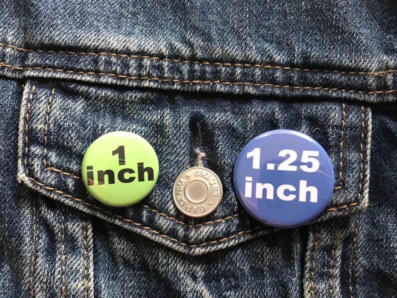 Peut inclure: Deux boutons ronds &eacute;pingl&eacute;s sur une veste en jean bleue. Le bouton de gauche est vert avec du texte noir qui indique "1 pouce". Le bouton de droite est bleu avec du texte blanc qui indique "1,25 pouce".