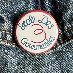 May include: A round button with the text "Ecole Des Gourmandes" in blue and red script. The button has a white background with a red border. The button is pinned to a blue denim jacket.