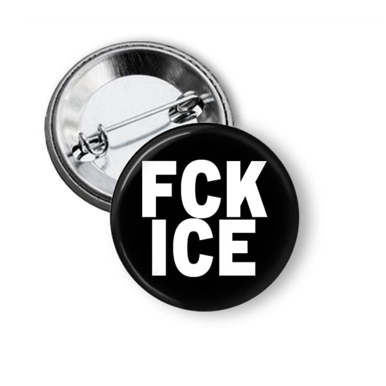 Puede incluir: Un bot&oacute;n negro con el texto blanco "FCK ICE". El bot&oacute;n es redondo y tiene un respaldo de alfiler met&aacute;lico. Otro bot&oacute;n es parcialmente visible en el fondo.