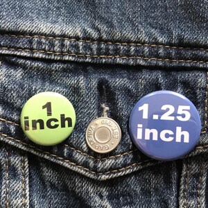 Puede incluir: Primer plano de un bolsillo de chaqueta vaquera con tres botones. Un bot&oacute;n verde claro que dice "1 inch" (2,54 cm), un bot&oacute;n plateado y un bot&oacute;n azul que dice "1.25 inch" (3,18 cm). Los botones son redondos y tienen texto negro.