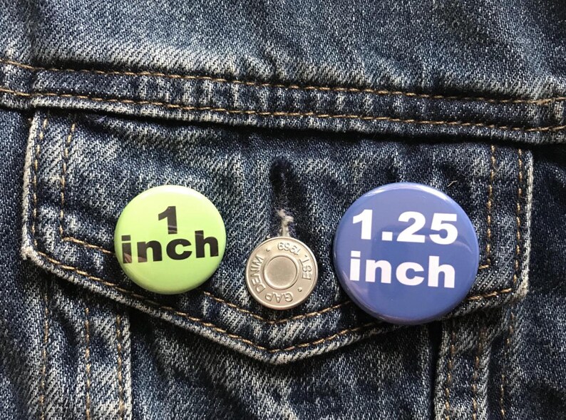 Puede incluir: Primer plano de un bolsillo de chaqueta vaquera con tres botones. Un bot&oacute;n verde claro que dice "1 inch" (2,54 cm), un bot&oacute;n plateado y un bot&oacute;n azul que dice "1.25 inch" (3,18 cm). Los botones son redondos y tienen texto negro.
