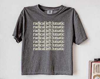 May include: Dark gray t-shirt with the text "radical left lunatic" repeated multiple times in a stacked layout. The shirt is hanging on a white wall, near a disco ball and a vintage radio.