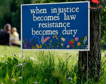 May include: A blue sign with white text that reads "when injustice becomes law resistance becomes duty." The sign has a floral design at the bottom and is in a grassy area. The Lucky Puppy Co. logo is in the upper right corner.