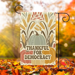 May include: A decorative garden flag with a fall harvest theme. The flag features a light green pumpkin, wheat stalks, and the text "THANKFUL FOR DEMOCRACY" in a stylized font. The flag is mounted on a black metal stand.