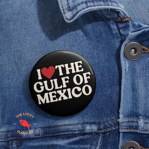 May include: A black button pin with the text "I ❤️ THE GULF OF MEXICO" in white, with a red heart symbol. The pin is on a denim jacket. The Lucky Guppy Co. logo is visible.