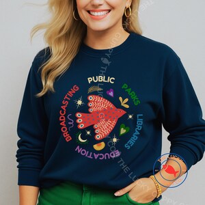 Könnte beinhalten: Marineblaues Sweatshirt mit einem kreisförmigen Design mit einem roten Vogel und den Worten "Public, Parks, Libraries, Education, Broadcasting". Das Design umfasst Herzen, Sterne und einen Mond. Das Sweatshirt hat einen Rundhalsausschnitt.