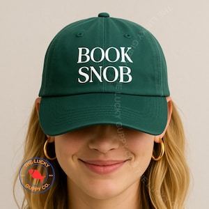 May include: A dark green baseball cap with the words "BOOK SNOB" in white block letters. The cap has a curved brim and a rounded top. The Lucky Guppy Co. logo is visible on the left side.