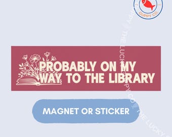 May include: A rectangular maroon sticker or magnet featuring the white text PROBABLY ON MY WAY TO THE LIBRARY. To the left of the text is a white line drawing of an open book with a bouquet of flowers emerging from the pages. The design is clean.