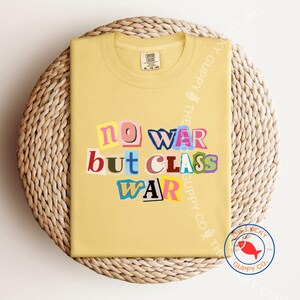 May include: A light yellow t-shirt with the text "NO WAR BUT CLASS WAR" in a colorful, cut-out style. The shirt is folded on a woven, circular surface. The Lucky Guppy Co. logo is visible in the bottom right corner.