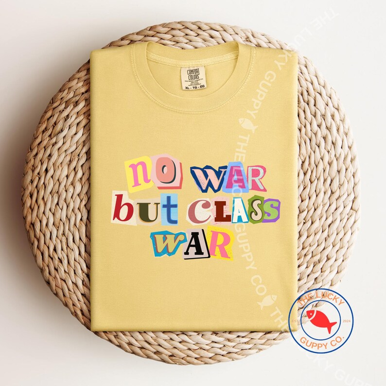 May include: A light yellow t-shirt with the text "NO WAR BUT CLASS WAR" in a colorful, cut-out style. The shirt is folded on a woven, circular surface. The Lucky Guppy Co. logo is visible in the bottom right corner.