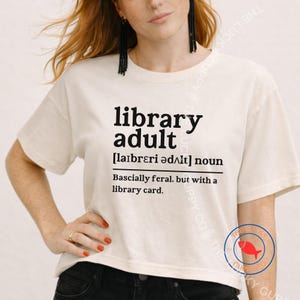 May include: Cream-colored t-shirt with the words "library adult" in black, along with a phonetic pronunciation and the phrase "Basically feral, but with a library card."