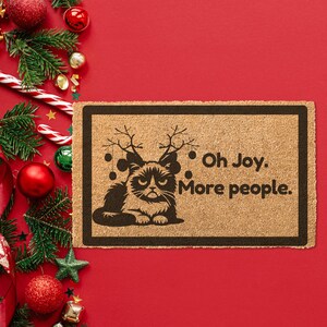 May include: A brown doormat with a black silhouette of a cat wearing reindeer antlers and the text "Oh Joy. More people."