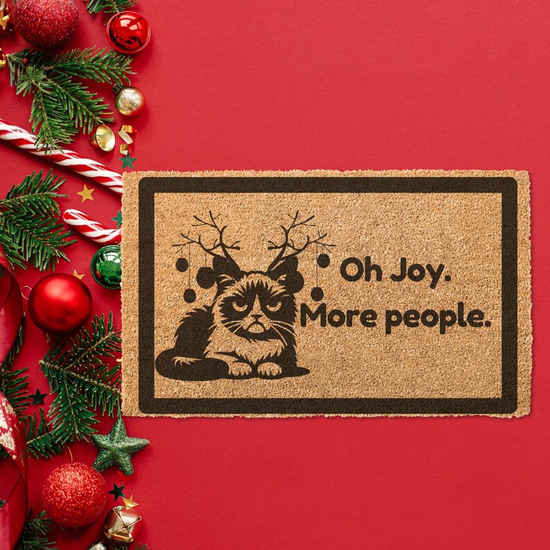 May include: A brown doormat with a black silhouette of a cat wearing reindeer antlers and the text "Oh Joy. More people."