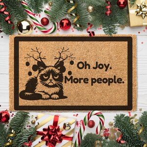 May include: A brown doormat with a black cat wearing reindeer antlers and the text "Oh Joy. More people."