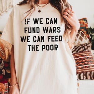 May include: Cream-colored t-shirt with the text "IF WE CAN FUND WARS WE CAN FEED THE POOR" in bold, black letters. The shirt has short sleeves and a relaxed fit. The image is taken in natural light.