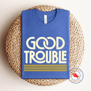 May include: A blue t-shirt with the words "GOOD TROUBLE" in a cream-colored font. Below the text are three horizontal yellow stripes. The shirt is displayed on a woven, circular surface.