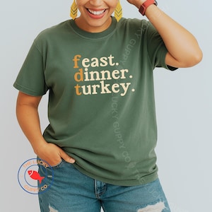 May include: Olive green t-shirt with the words "feast. dinner. turkey." in orange and tan lettering. The shirt has a crew neck and short sleeves. A circular logo with a red fish is visible on the bottom left.